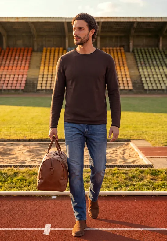 Mann in Pullover und Jeans mit Reisetasche geht auf einer Laufbahn im Stadion.