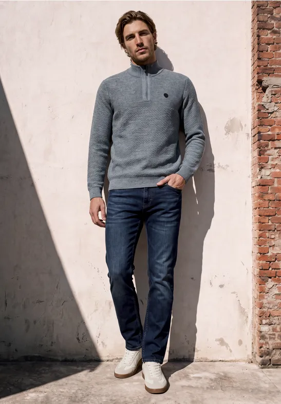 Ein Mann lehnt entspannt an einer sonnenbeschienenen Wand, trägt grauen Pullover, blaue Jeans und helle Sneaker.