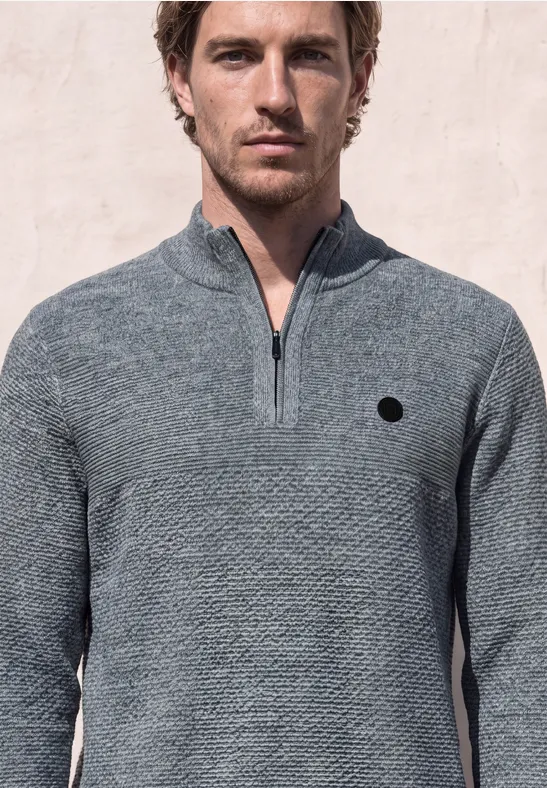 Ein Mann trägt einen grauen, texturierten Pullover mit halbem Reißverschluss vor hellem Hintergrund.