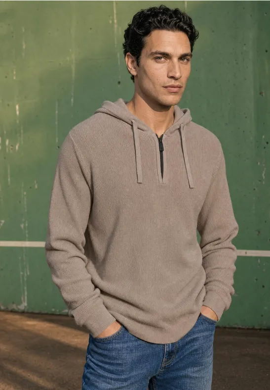 Ein junger Mann trägt einen beigen Hoodie und blaue Jeans vor grünem Hintergrund.
