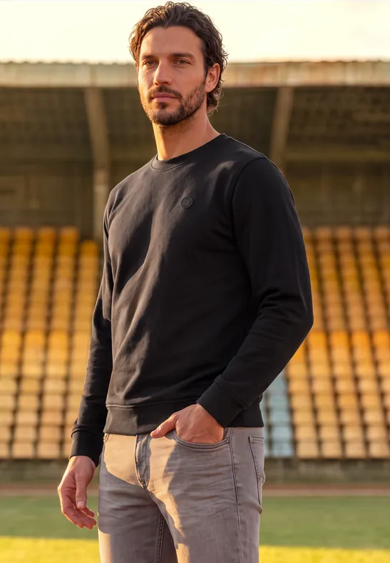 Ein bärtiger Mann im schwarzen Pullover steht vor gelben Tribünenplätzen im Stadion.