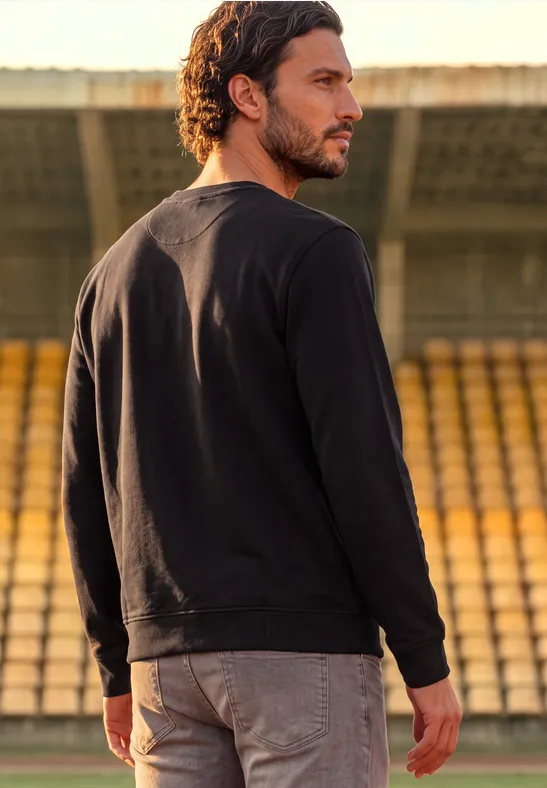 Ein Mann im schwarzen Pullover steht auf einer Tribüne und blickt in die Ferne.