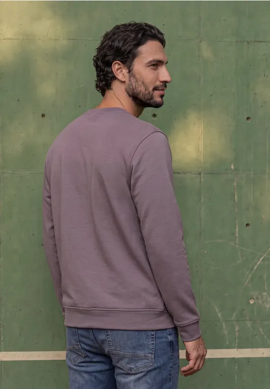 Ein Mann in lila Pullover und Jeans steht vor einer grünen Wand, Rückenansicht, dreht den Kopf seitlich.