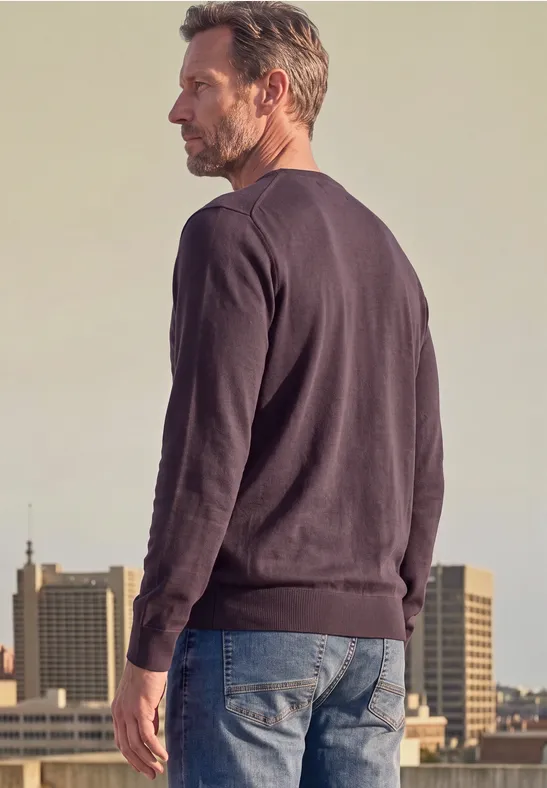 Ein Mann steht in Jeans und Pullover mit dem Rücken zur Kamera vor städtischer Kulisse.