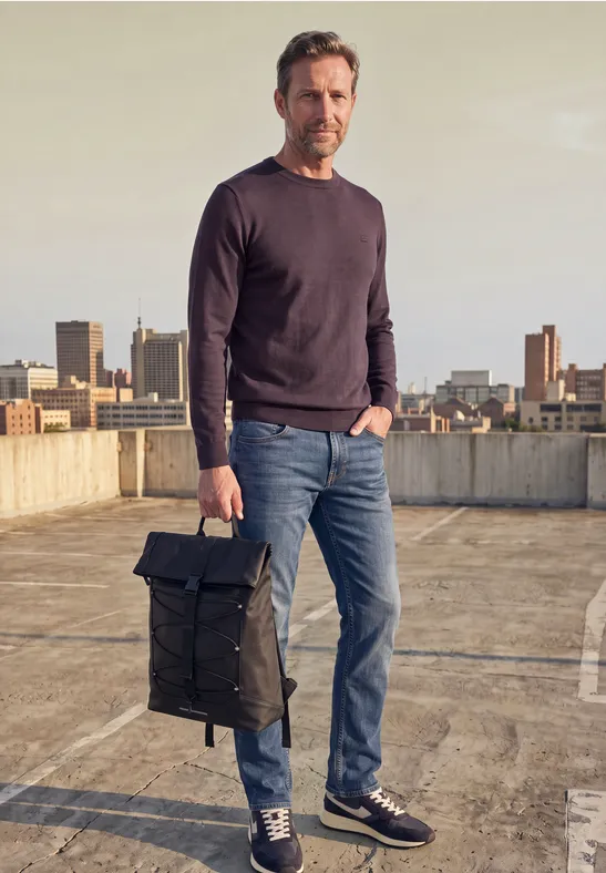 Ein Mann steht auf einem Parkdeck, trägt Jeans, Pullover und Sneaker und hält einen schwarzen Rucksack.