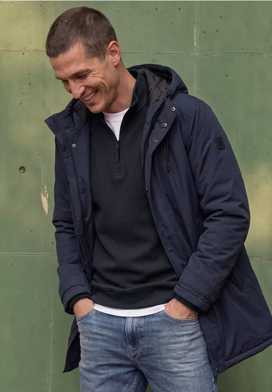 Mann in Jeans, Pullover und Kapuzenjacke lehnt lächelnd an grüner Wand.