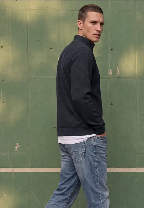Ein Mann in Jeans und Pullover steht vor einer grünen Wand und schaut zurück.