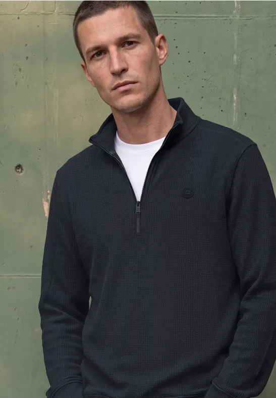 Ein Mann vor grünem Metallhintergrund trägt einen dunkelblauen Half-Zip-Pullover und ein weißes T-Shirt.