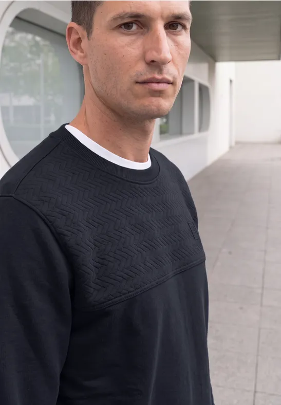 Ein Mann im schwarzen Pullover mit geometrischem Muster steht vor modernem Gebäude.