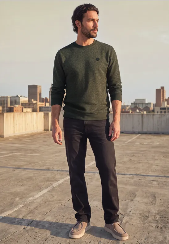 Mann in grünem Pullover und dunklen Hosen steht auf Parkdeckdach mit Stadtblick, schaut seitlich.