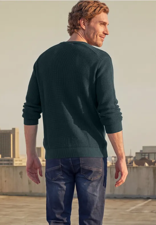 Un homme en pull vert et jean, de dos, se tient sur un toit avec vue urbaine.