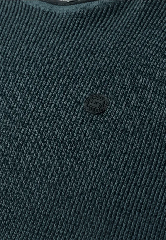 Gros plan d’un pull en maille gris foncé à col en V avec un petit écusson-logo discret sur la poitrine.