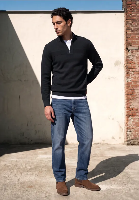 Ein Mann mit schwarzem Pullover, Jeans und braunen Schuhen steht draußen an einer Wand und schaut nach unten.