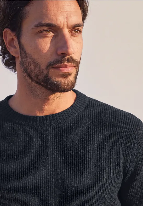 Portret van een peinzende man met donkere trui, baard en licht golvend haar.