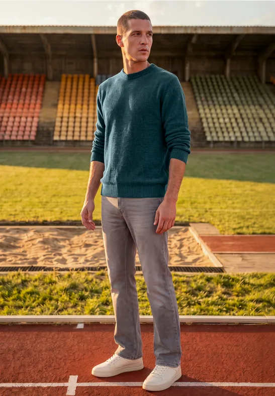 Ein Mann im Pullover und Jeans steht auf einer Laufbahn vor leeren Stadionrängen und schaut zur Seite.