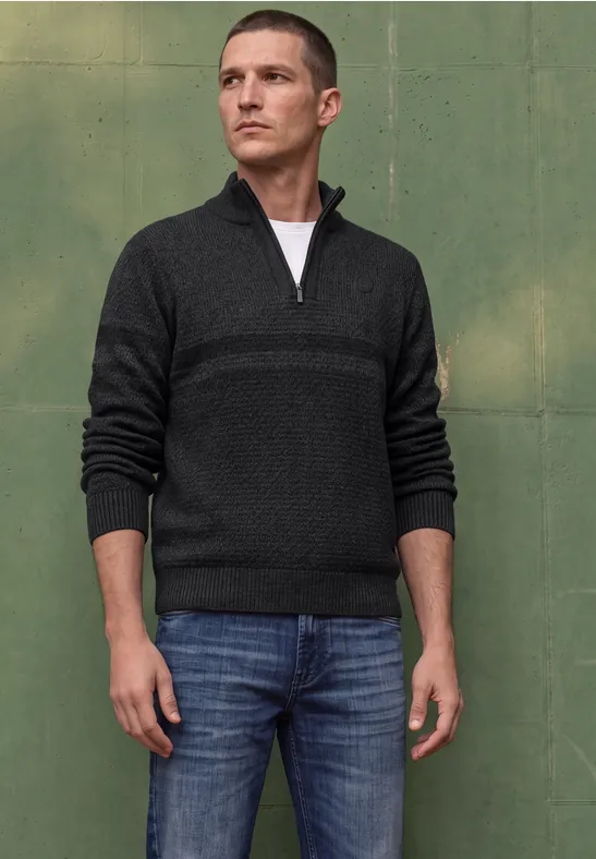 Ein Mann in dunkelgrauem Pullover mit Reißverschluss und Jeans vor grünem Hintergrund.