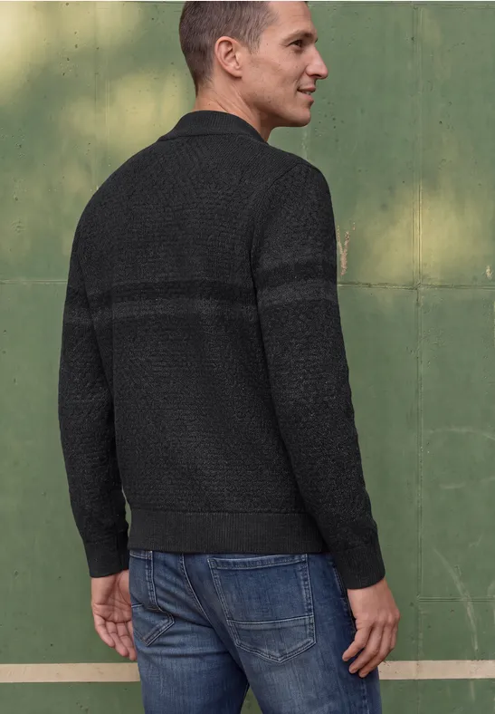 Ein Mann steht mit dem Rücken zur Kamera, trägt schwarzen Pullover und blaue Jeans vor grüner Wand.