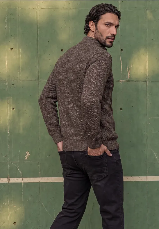 Ein Mann in braunem Pullover und schwarzer Hose steht seitlich vor einem grünen Tennisplatz und blickt zurück.
