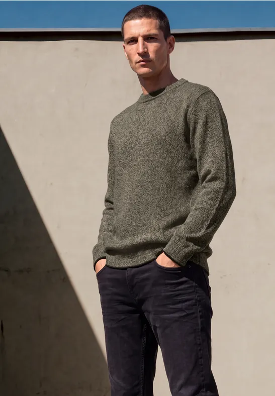 Ein Mann trägt einen grünen Strickpullover und schwarze Hose vor einer hellen Wand.