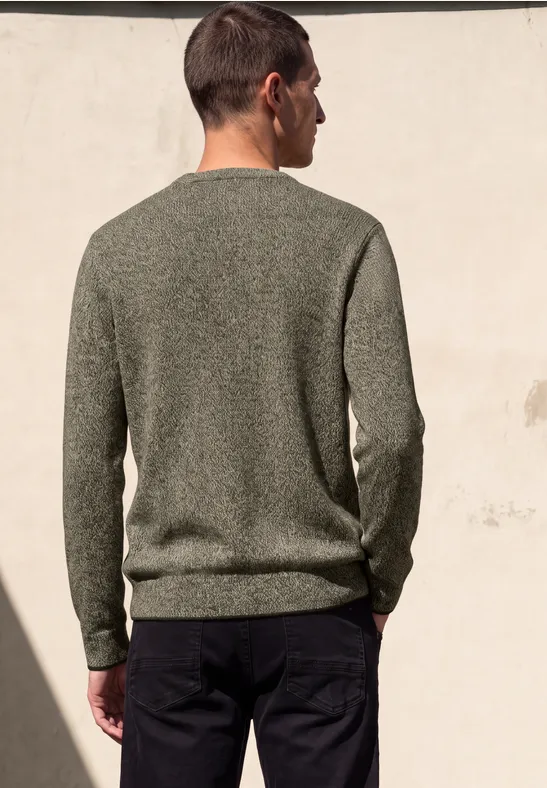 Ein Mann steht mit dem Rücken zur Kamera, trägt einen grünen Pullover und dunkle Hose vor heller Wand.