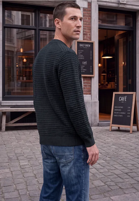 Ein Mann in Jeans und Pullover blickt über die Schulter auf einen gepflasterten Weg vor einem Café.