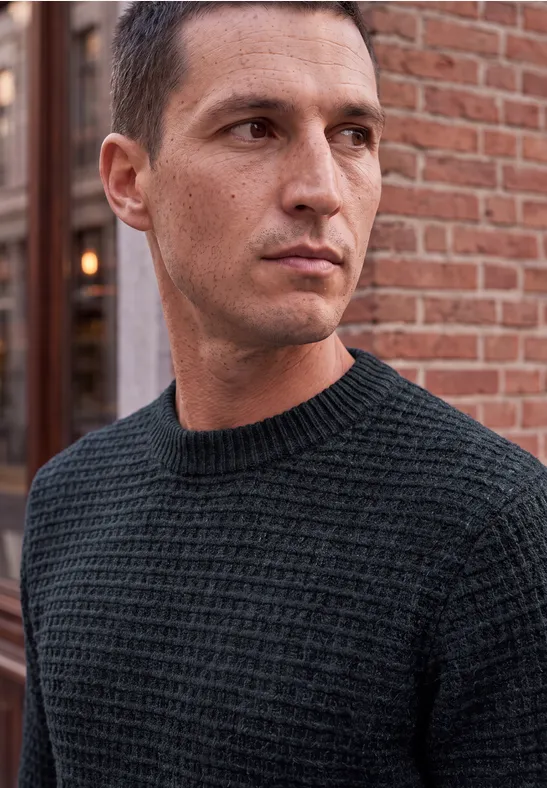 Ein Mann mit kurzem dunklem Haar und schwarzem Strickpullover schaut seitlich vor einer Backsteinwand.