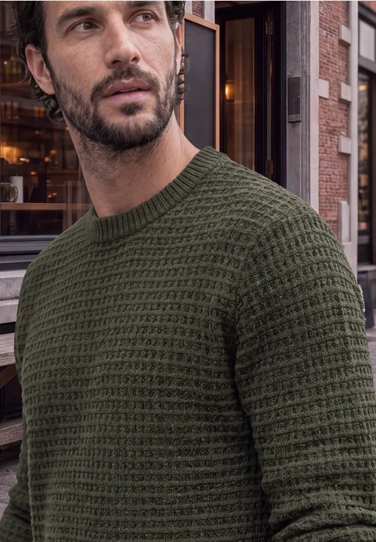 Mann mit olivgrünem, grob gestricktem Pullover vor städtischem Gebäude