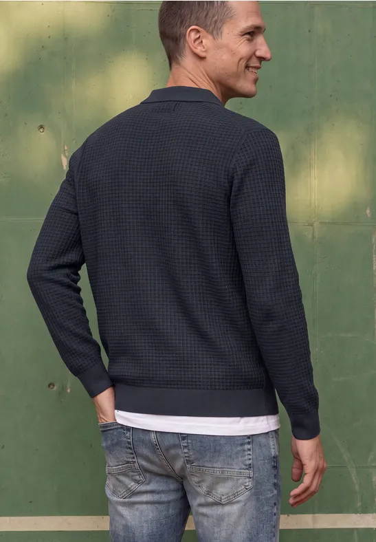 Ein Mann steht seitlich, trägt dunkelblauen Pullover, Jeans, sieht nach links, grüner Hintergrund.