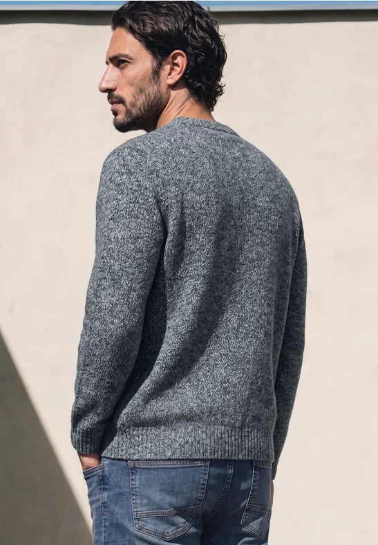 Ein Mann trägt einen grauen Pullover und Jeans, steht seitlich vor einer hellen Wand.