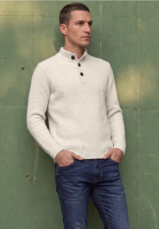 Mann lehnt vor grünem Hintergrund, trägt hellen Pullover und Jeans.