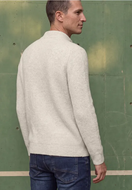Ein Mann in hellem Pullover und Jeans steht mit dem Rücken zur Kamera vor einer grünen Wand.