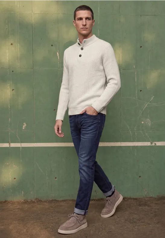 Ein Mann in Jeans, weißem Pullover und grauen Sneakern vor grüner Wand mit weißer Linie.