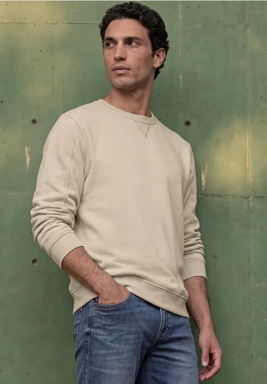 Ein Mann in hellem Pullover und Jeans lehnt an einer grünen Wand und blickt zur Seite.