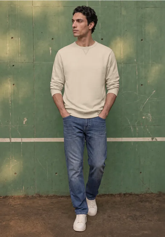 Ein Mann steht vor einer grünen Wand, trägt helles Langarmshirt, blaue Jeans und weiße Sneaker, Hände in den Taschen.