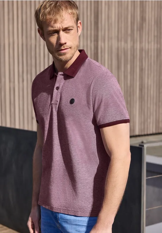 Ein Mann in pinker Polo-Shirt steht vor einer Holzwand und blickt zur Seite.