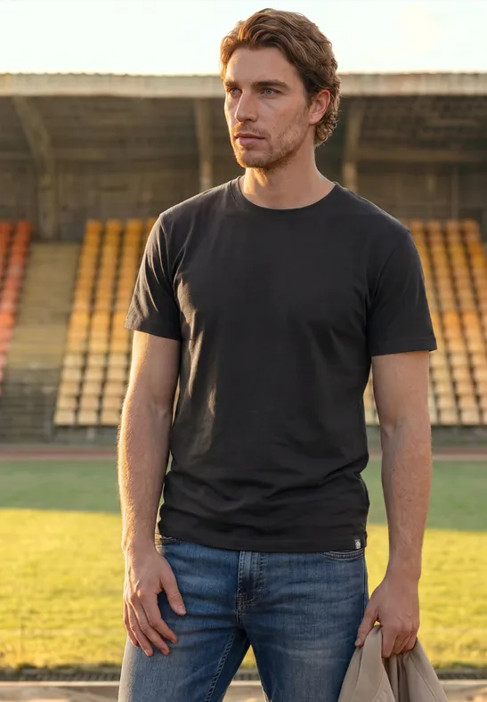 Ein Mann in schwarzem T-Shirt und Jeans steht auf dem Rasen vor leeren Stadionrängen und blickt zur Seite.