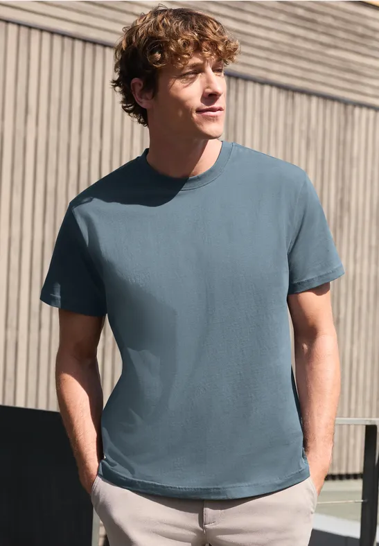 Ein Mann steht vor einer Hauswand und trägt lässig ein schlichtes blaues T-Shirt.