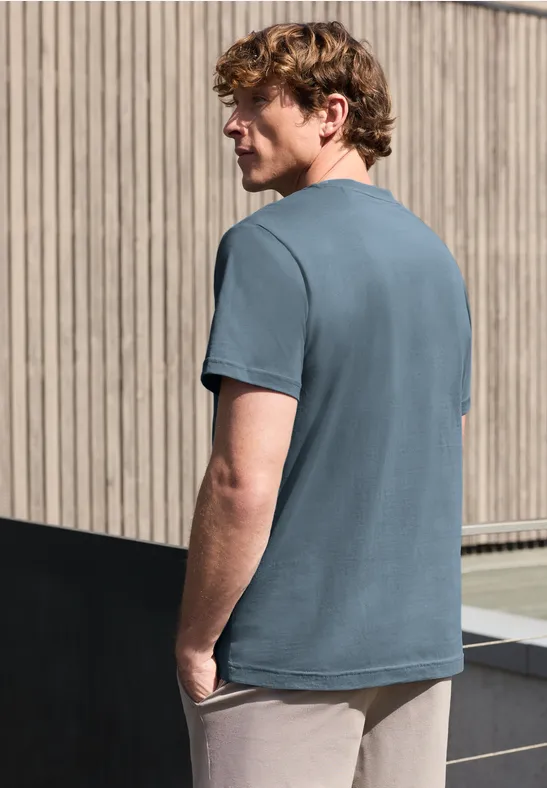 Ein Mann steht mit Rücken zum Betrachter, trägt ein graublaues T-Shirt vor einer Holzfassade.