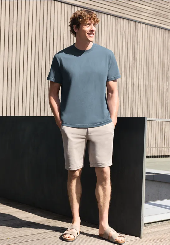 Ein junger Mann trägt ein blaues T-Shirt, beige Shorts und Sandalen, steht lässig vor einer Holzfassade.