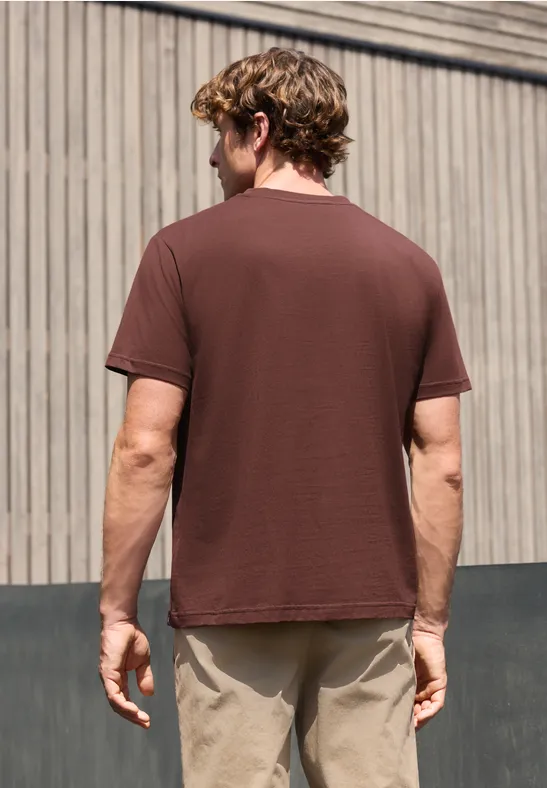 Ein Mann von hinten vor Holzfassade, braunes T-Shirt und beige Hose