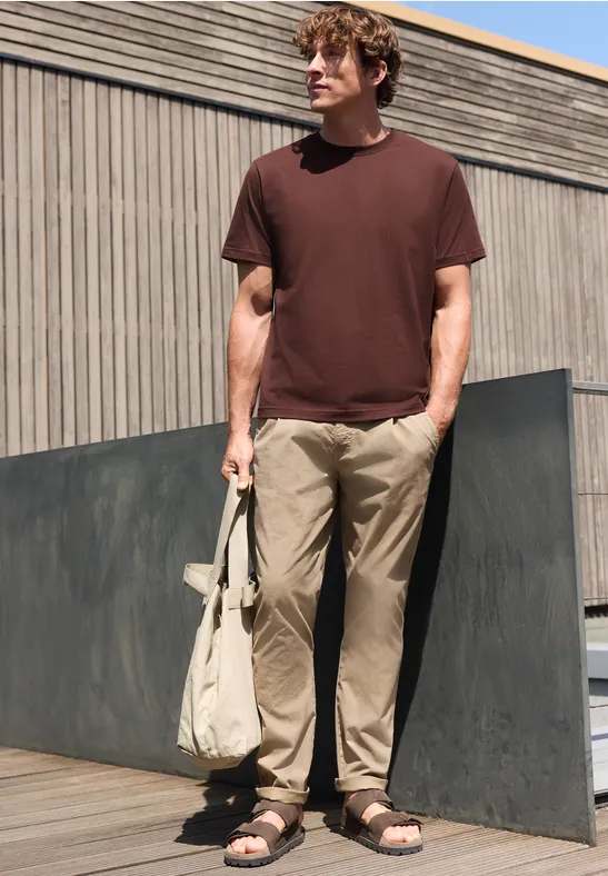Ein Mann steht draußen vor einer Holzfassade, trägt braunes T-Shirt, helle Hose und Sandalen, hält eine Tasche.