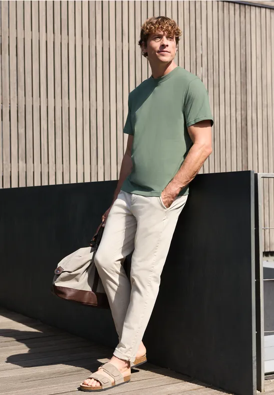 Ein Mann lehnt an einer Wand, trägt ein grünes T-Shirt, helle Hose, Sandalen und hält eine Reisetasche.