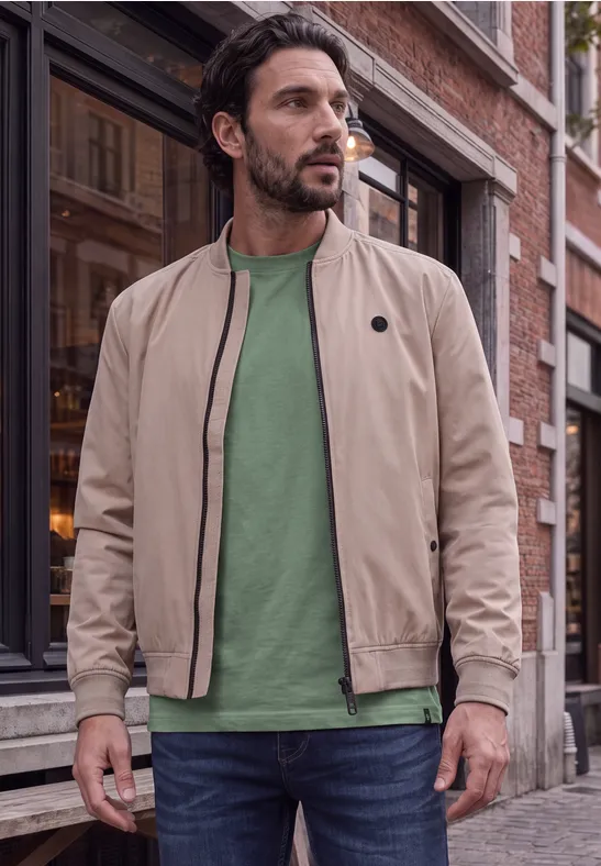 Ein Mann trägt eine beige Bomberjacke und ein grünes Shirt und steht vor einem Backsteingebäude.