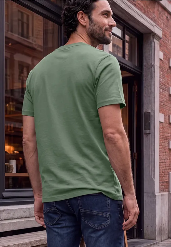Ein Mann steht mit dem Rücken zur Kamera, trägt ein grünes T-Shirt und Jeans vor einem Gebäude.
