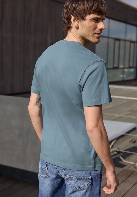 Ein Mann steht auf einer Terrasse und blickt über die Schulter, trägt ein türkis-blaues T-Shirt und Jeans.