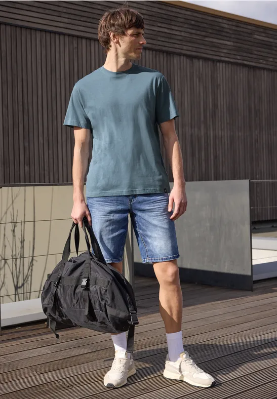 Ein Mann steht auf einer Holzterrasse, trägt blauen T-Shirt, Jeans-Shorts, weiße Sneaker und hält eine Tasche.