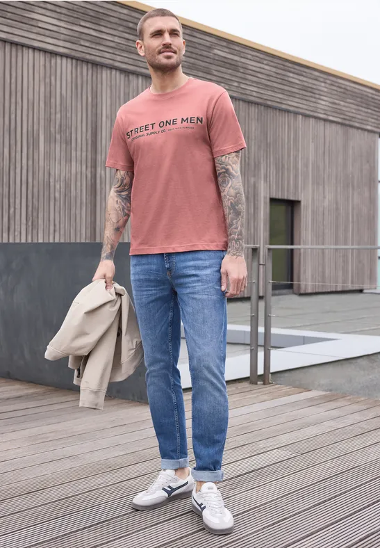 Ein tätowierter Mann in lachsfarbenem T-Shirt, blauen Jeans und weißen Sneakern hält eine Jacke.