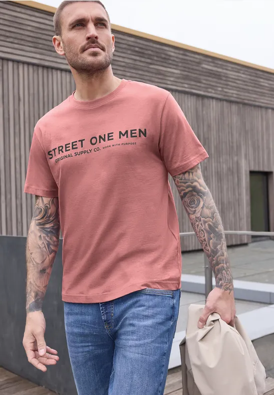 Ein tätowierter Mann trägt ein rosafarbenes T-Shirt mit 'STREET ONE MEN', Jeans und hält eine Tasche.