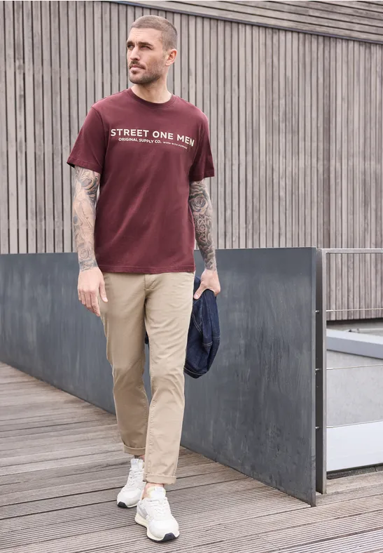 Ein tätowierter Mann trägt ein bordeauxfarbenes T-Shirt, beigen Chinos und weiße Sneaker und hält eine Jacke.