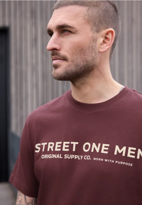 Junger Mann trägt ein dunkelrotes T-Shirt mit weißer Aufschrift 'STREET ONE MEN' vor neutralem Hintergrund.